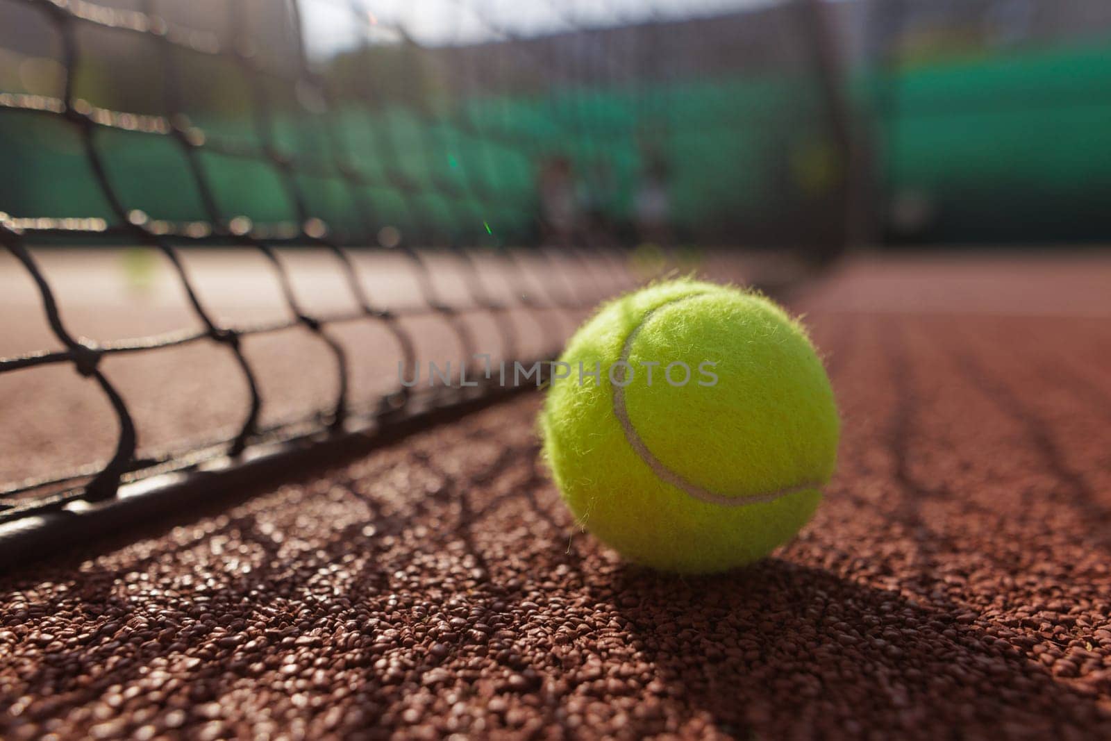 A close-up of a tennis ball resting on a clay court, with a net in the foreground and blurred players in the background