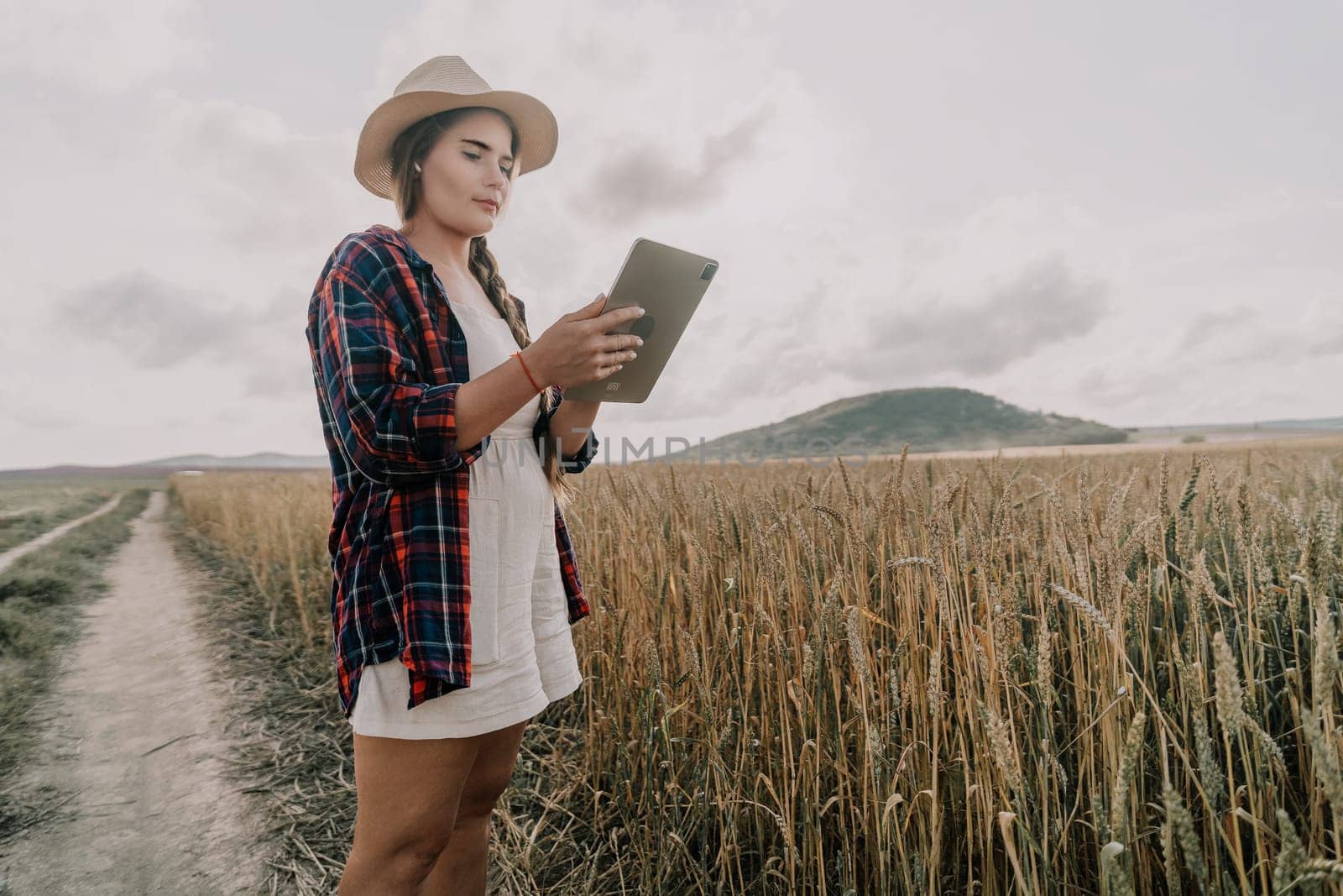 A young woman in a plaid shirt and straw hat uses her tablet while standing in a wheat field.