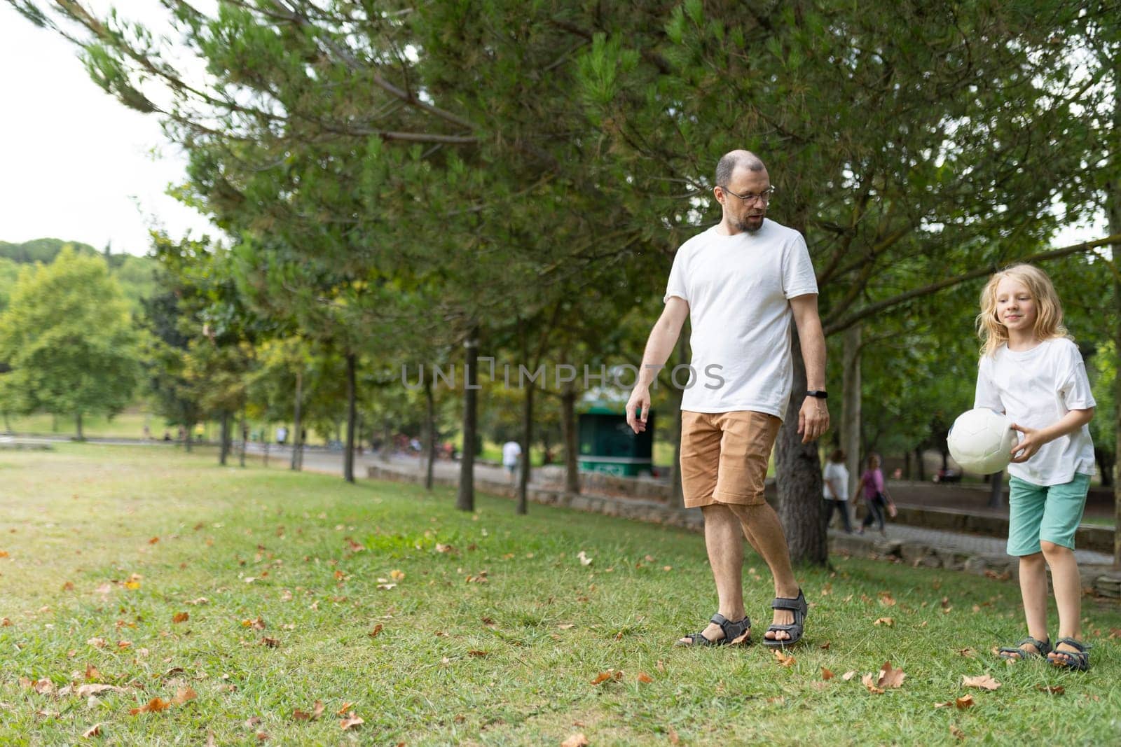Father and daughter are enjoying a leisurely stroll through a lush green park, carrying a volleyball on a sunny day