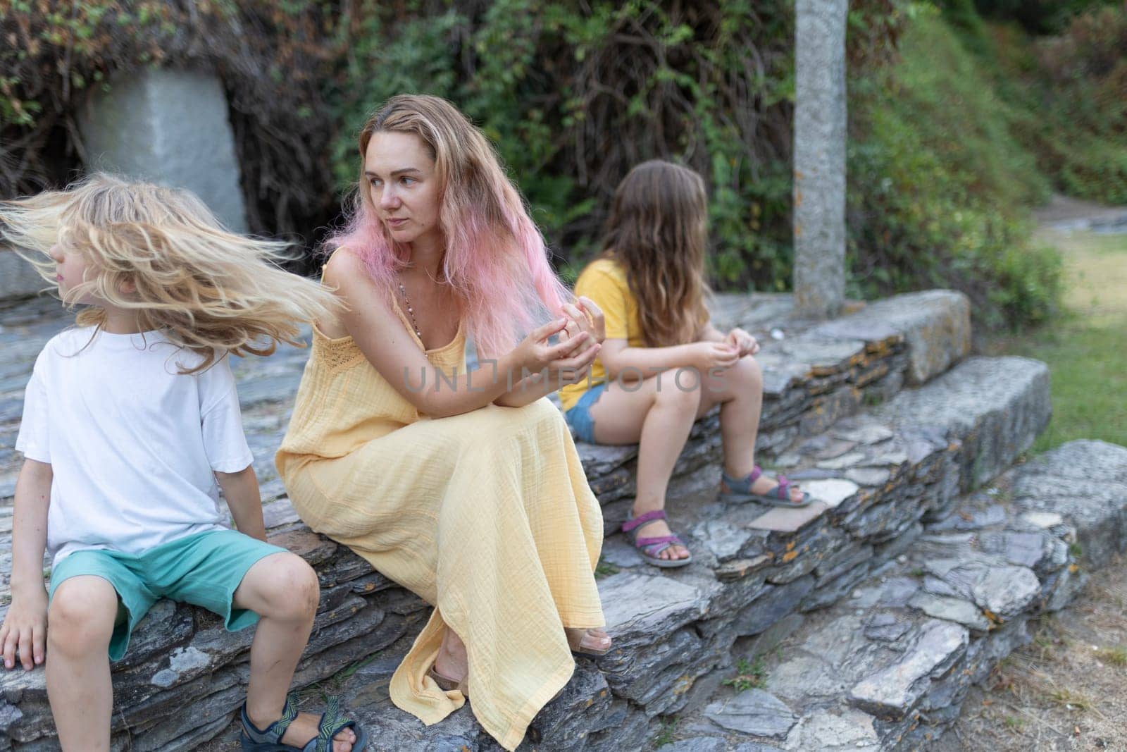 Mother with pink hair wearing a yellow dress is sitting on stone steps with her two daughters, seemingly sad and thoughtful
