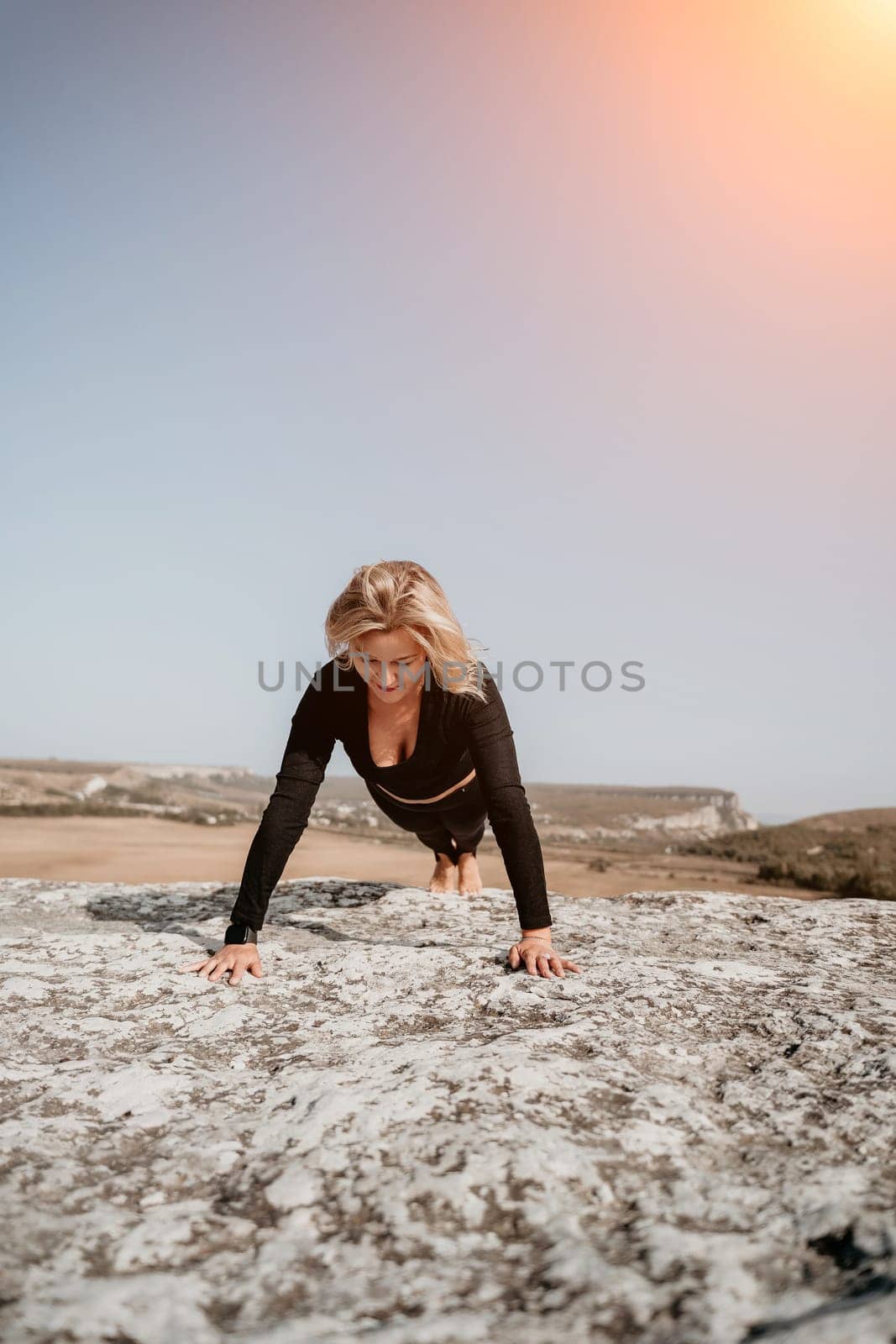 A woman in black athletic wear does push ups on a rocky cliff with a scenic view behind her.