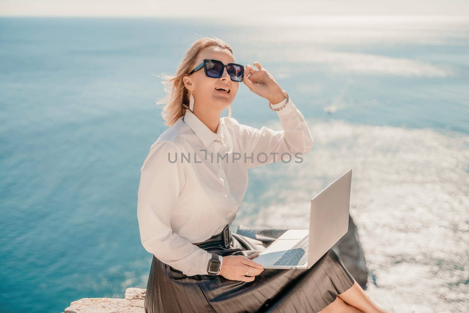 Business woman on nature in white shirt and black skirt. She works with an iPad in the open air with a beautiful view of the sea. The concept of remote work