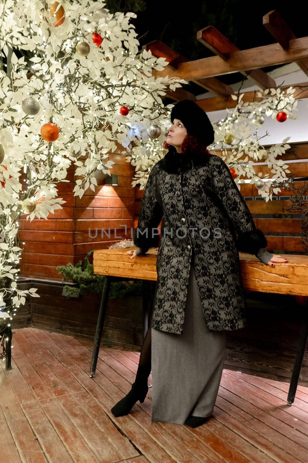 A woman in a black coat stands in front of a Christmas tree. She is wearing a hat and a scarf