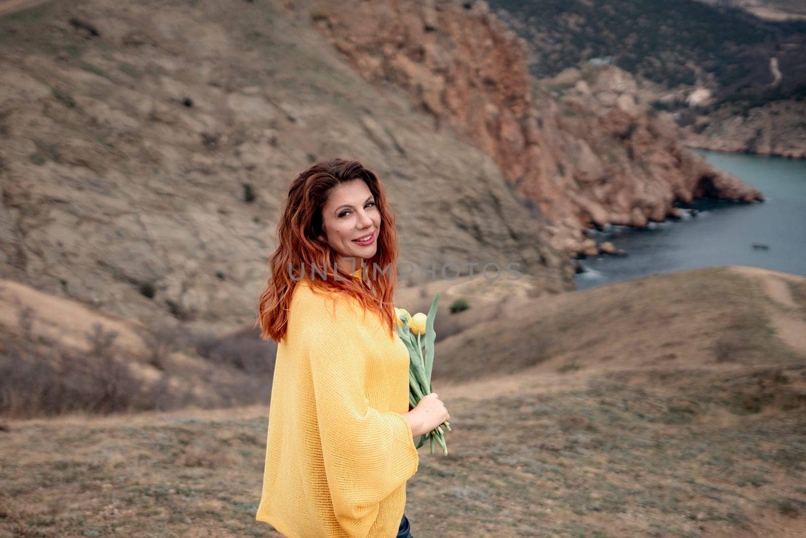 Portrait of a happy woman with long hair against a background of mountains and sea. Holding a bouquet of yellow tulips in her hands, wearing a yellow sweater.
