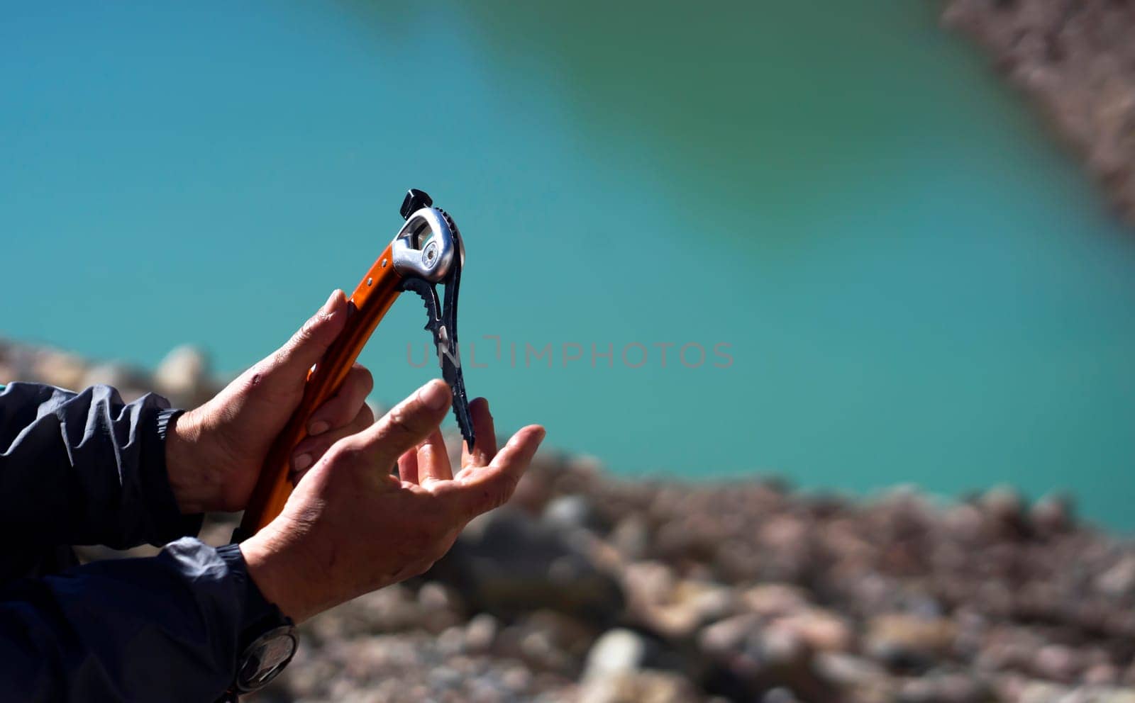 Male hands hold an ice ax on the background of a mountain lake from a glacier. A man takes ice tools while climbing the mountain.