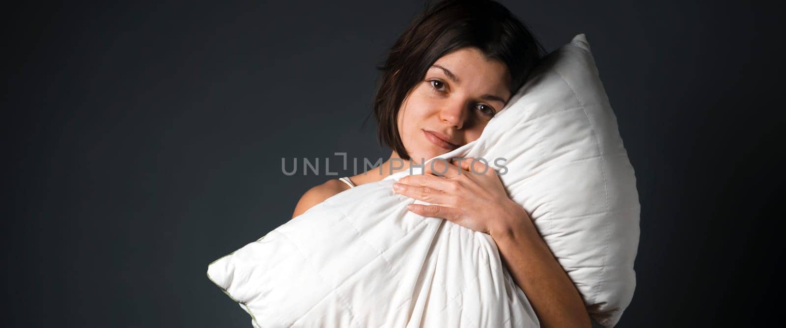 A young girl stands and hugs a white pillow on a gray background, a woman wants to sleep, take care of her health, the quality of sleep.