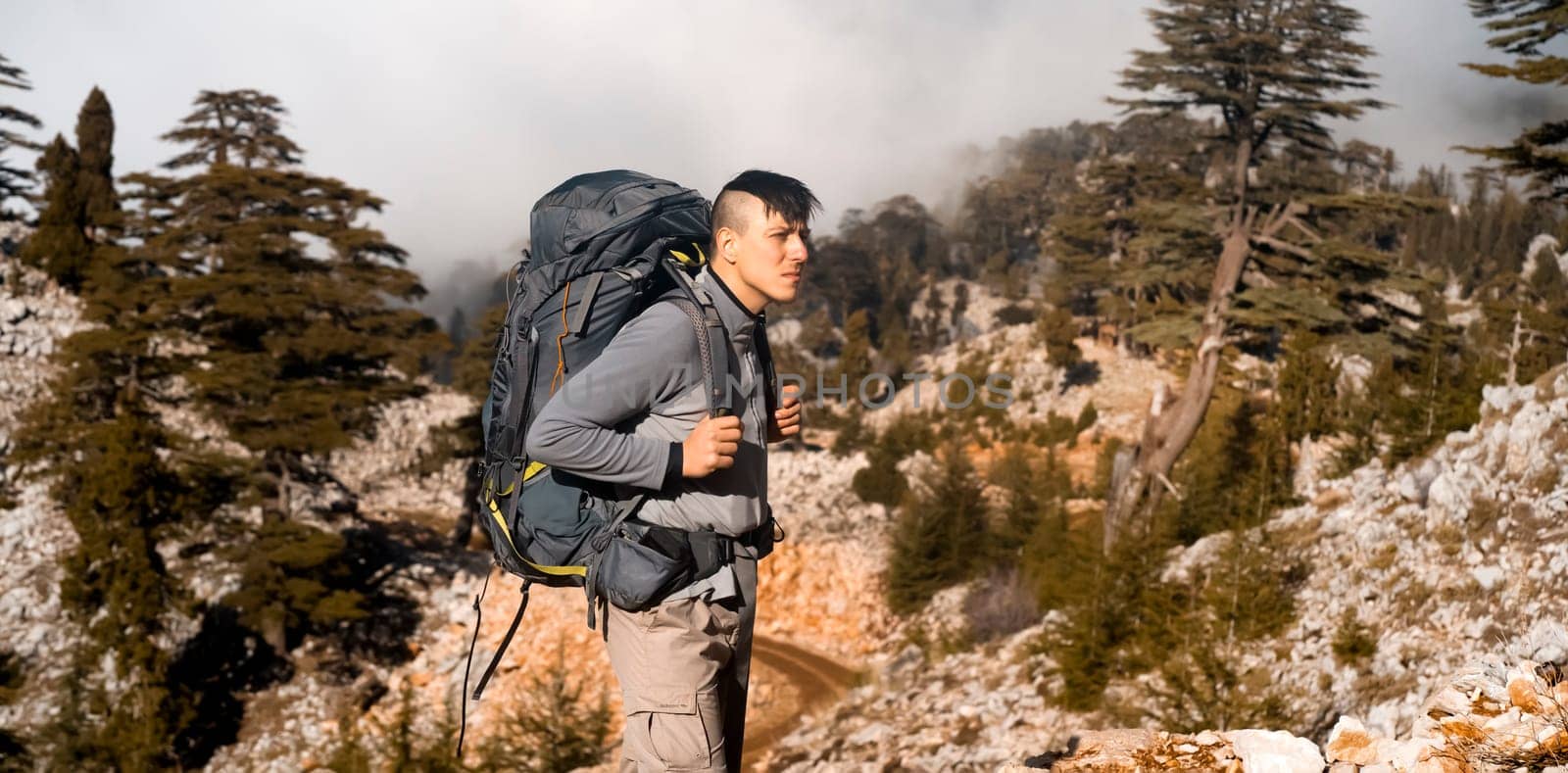 A young man with tourist equipment in a backpack, in trekking pants and a fleece jacket climbs to the top of the mountain. The traveler goes hiking in early spring. Rocks and stones on the background.