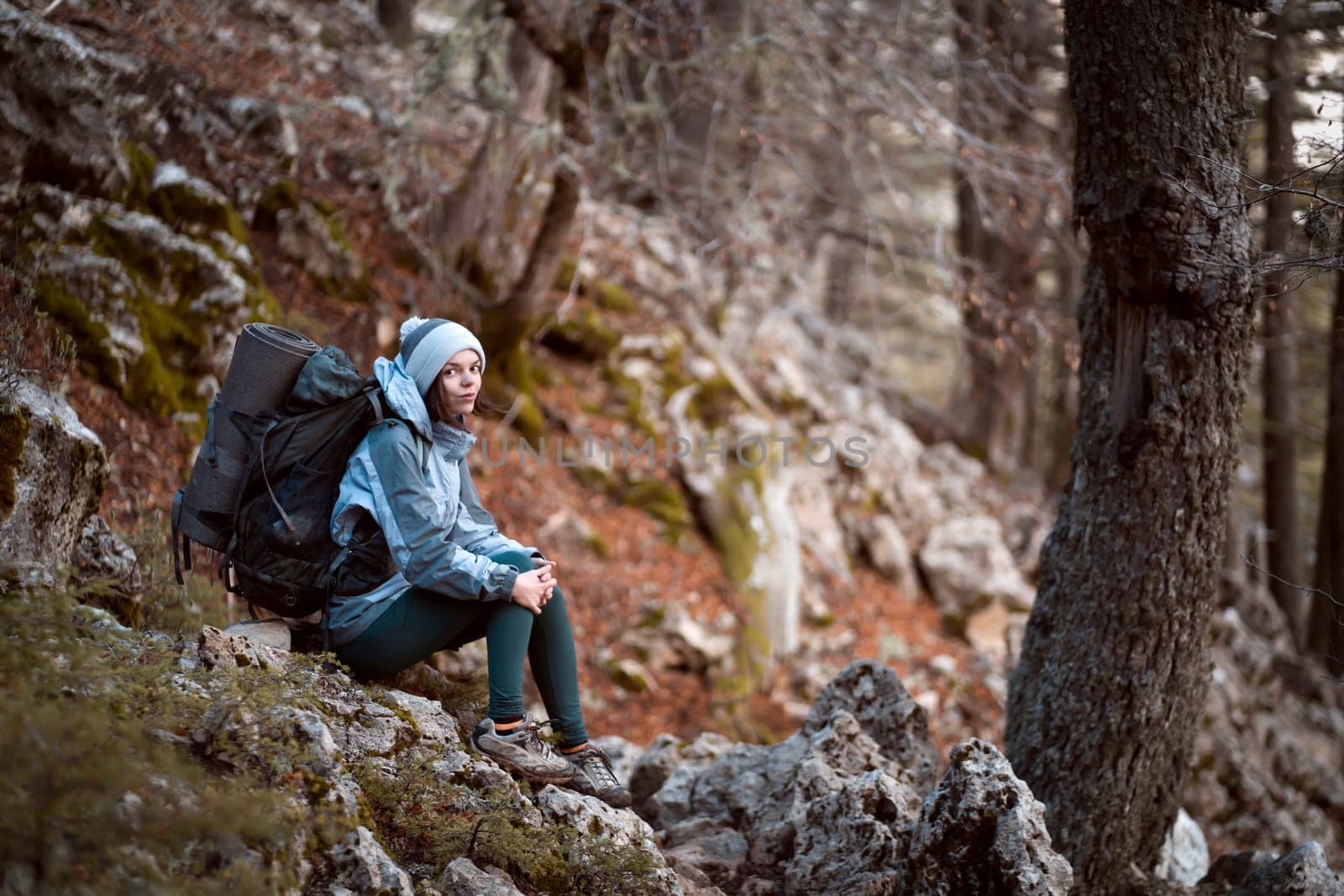 A young girl sits with a backpack and hiking equipment on a stone, rests during her climbing in the mountains and forest, the woman leads an active lifestyle and is involved in mountaineering.