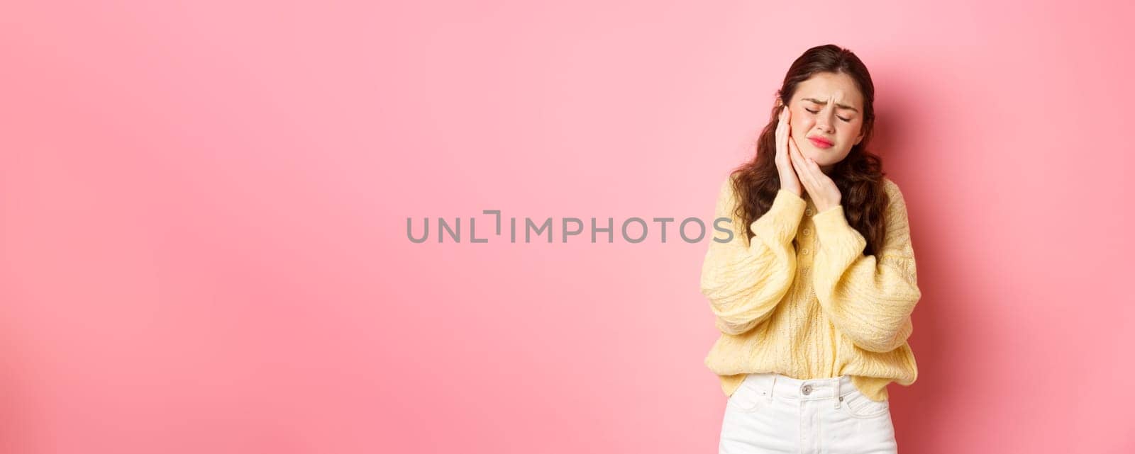 Young woman with terrible toothache, touching swallen cheek and grimacing from painful feeling, having tooth decay, need appointment to dentist, standing against pink background.