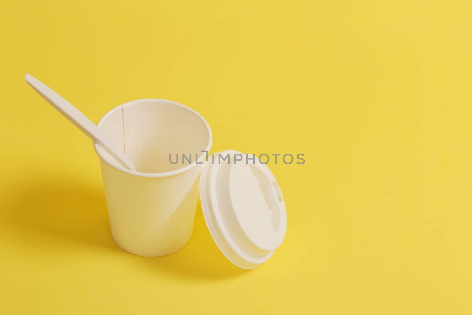 Disposable glass with a lid, with a teaspoon on a yellow background.