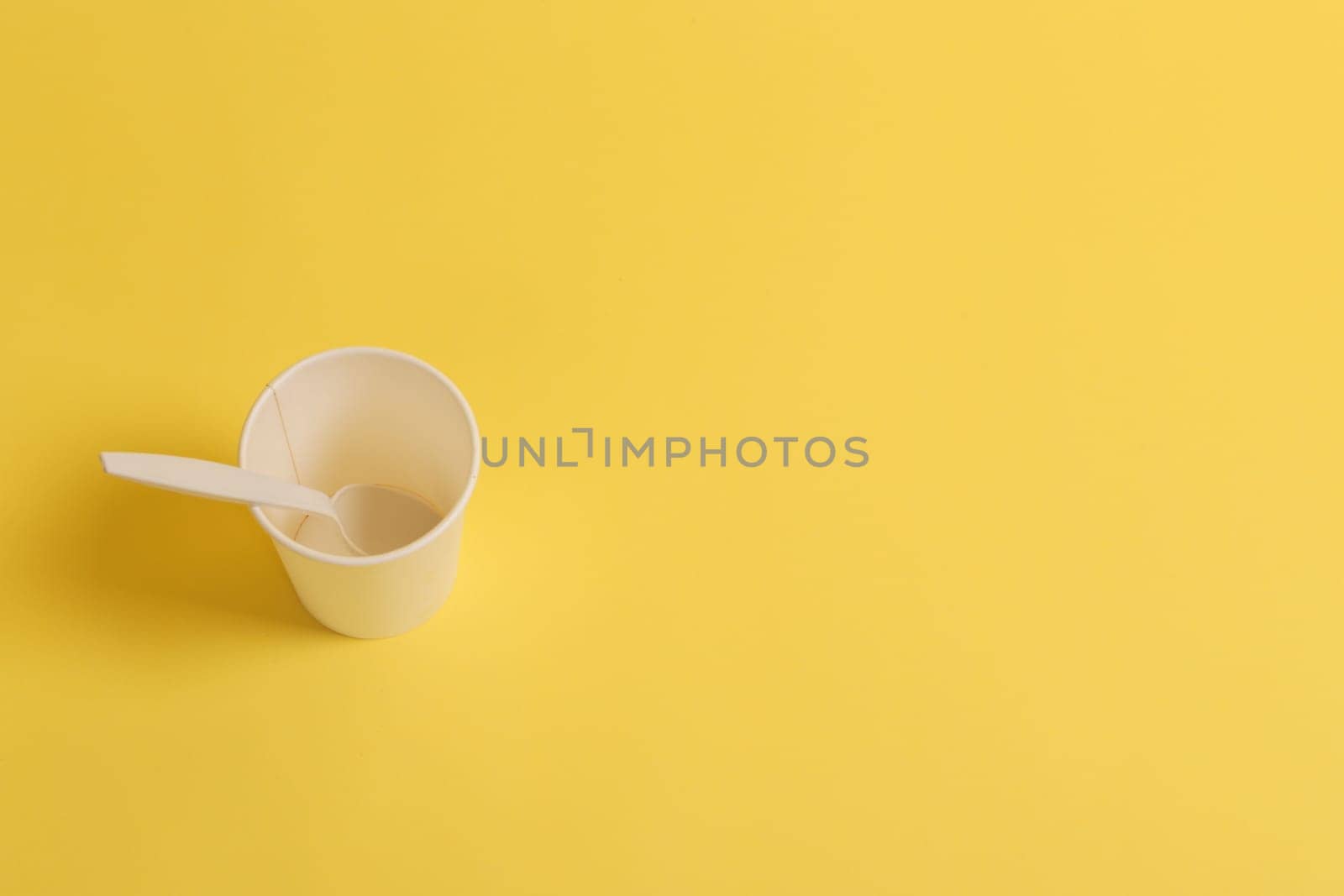 Paper glass with a plastic spoon on a yellow background, top view.