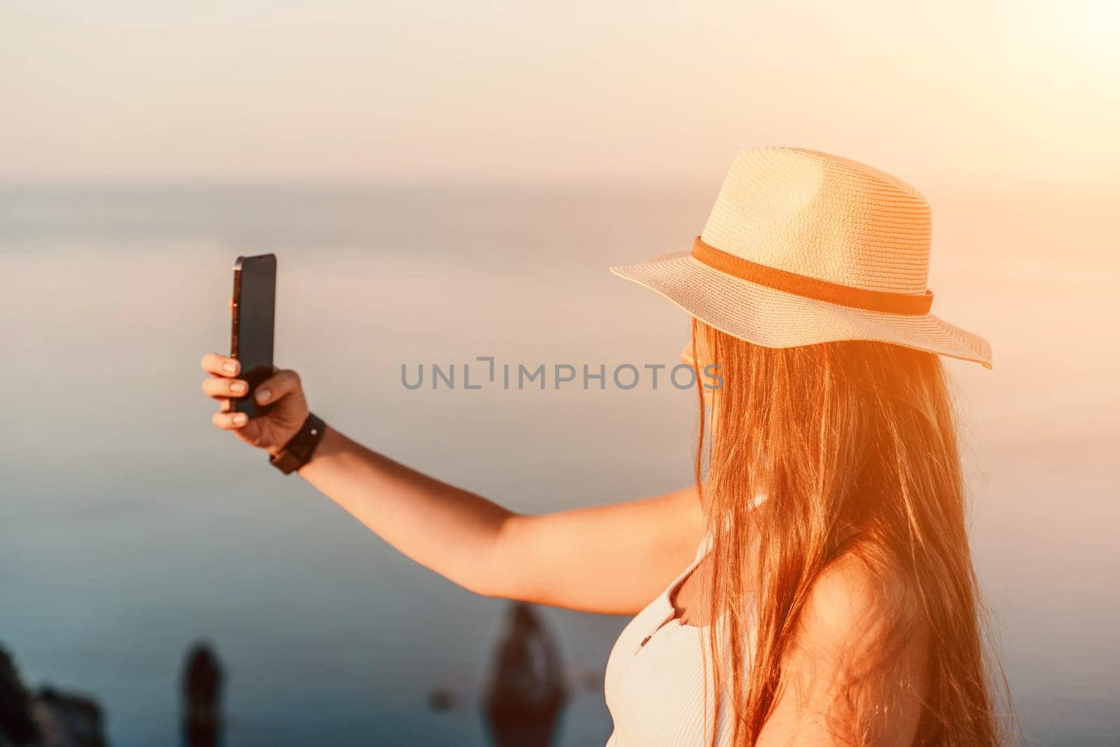 Selfie woman in a hat, white tank top, and shorts captures a selfie shot with her mobile phone against the backdrop of a serene beach and blue sea