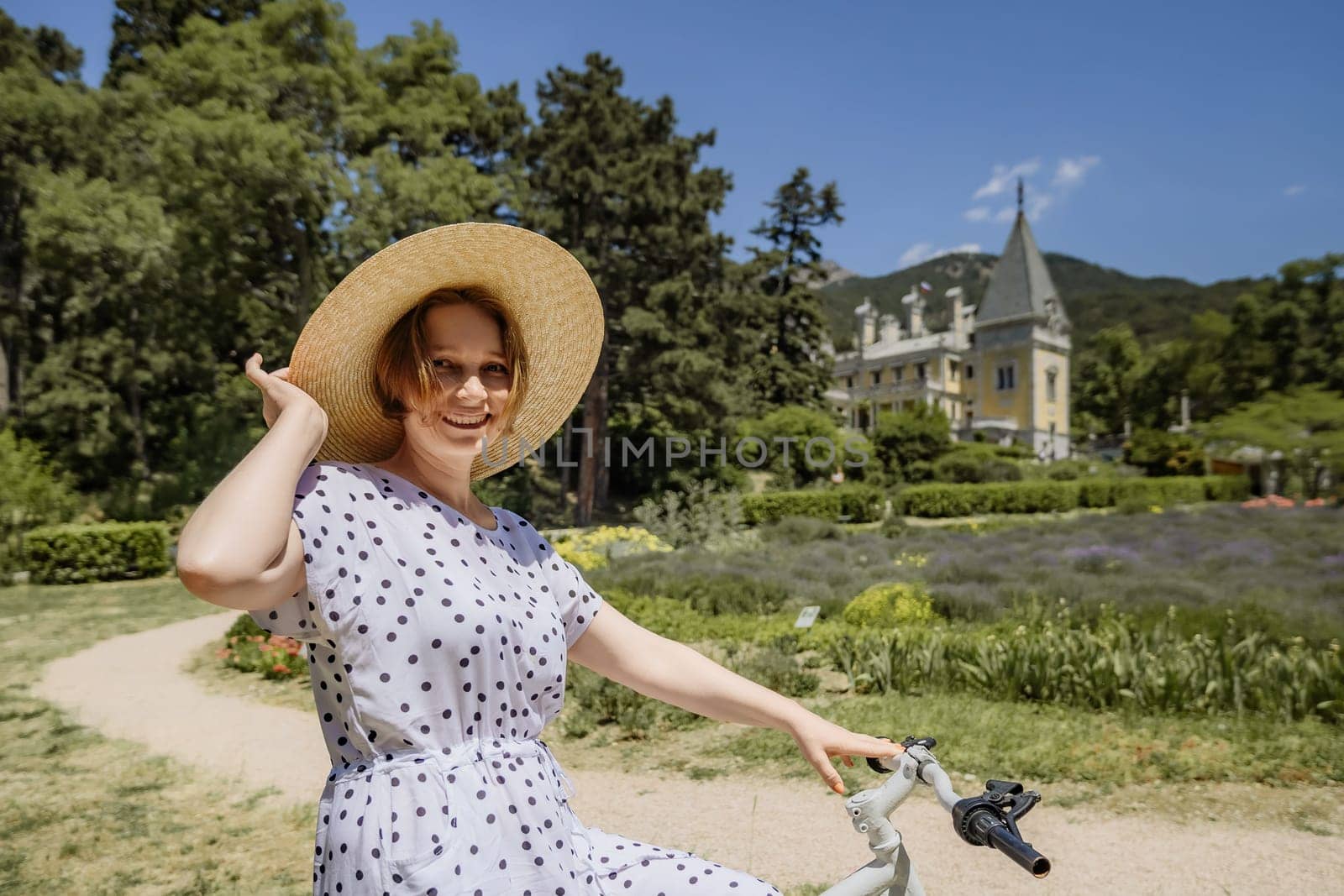 A woman wearing a straw hat and a white polka dot dress is riding a bicycle in a park