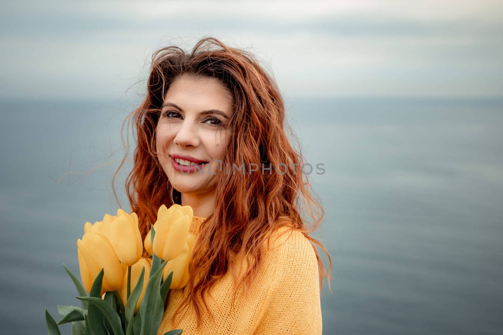 Portrait of a happy woman with hair flying in the wind against the backdrop of mountains and sea. Holding a bouquet of yellow tulips in her hands, wearing a yellow sweater.
