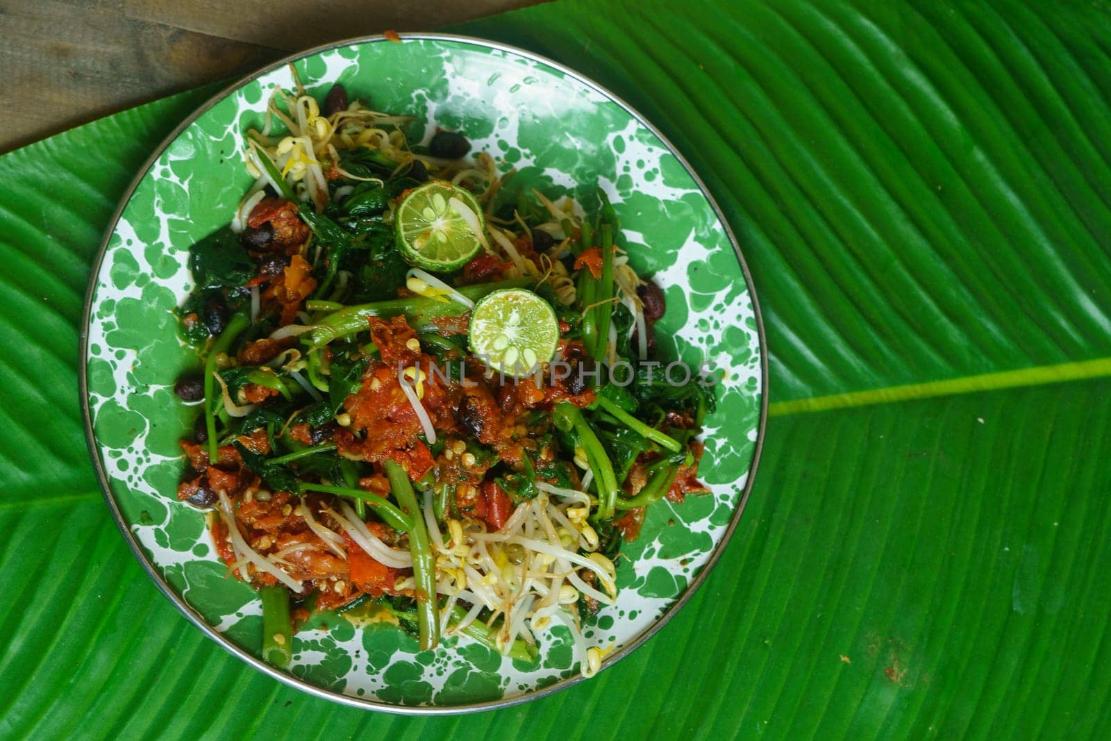 A vibrant plate of Plecing Kangkung, Lombok, Indonesian traditional food. Featuring stir fried water spinach topped with a spicy sambal made from chili peppers, tomatoes, shallots, and garlic, garnished with bean sprouts and lime wedges.
