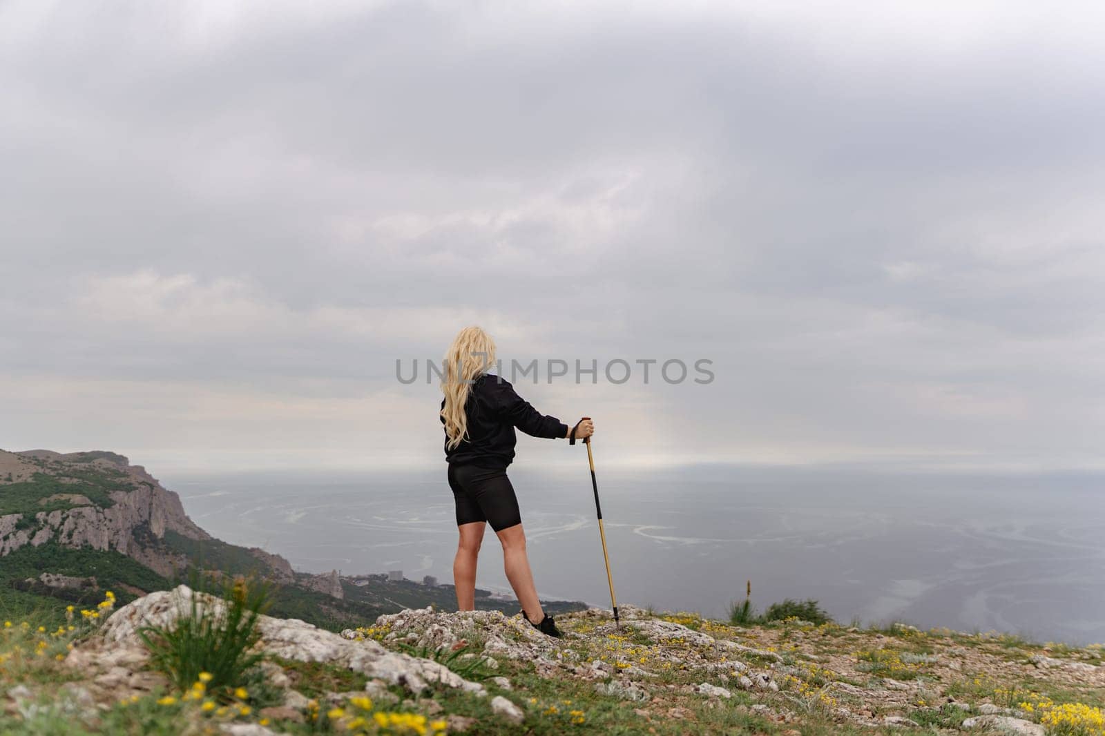 A woman stands on a rocky hill overlooking the ocean. She is holding a walking stick and wearing shorts. The sky is cloudy, and the ocean is visible in the distance