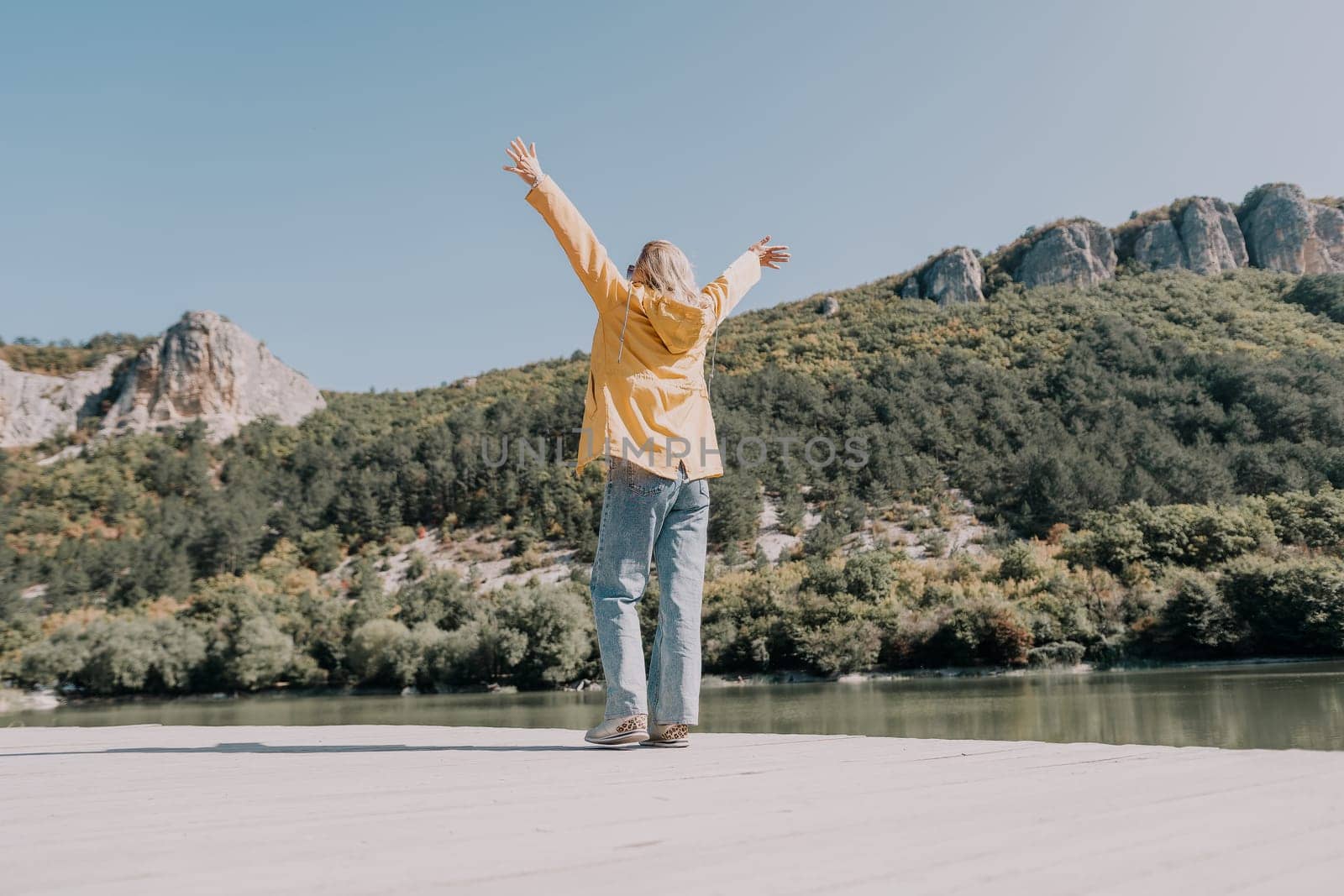 A young woman in a yellow jacket stands on a wooden dock, arms outstretched towards the mountains and lake. She is wearing blue jeans and sneakers, facing a beautiful natural scenery. The sunny, calm atmosphere conveys a sense of freedom and joy.