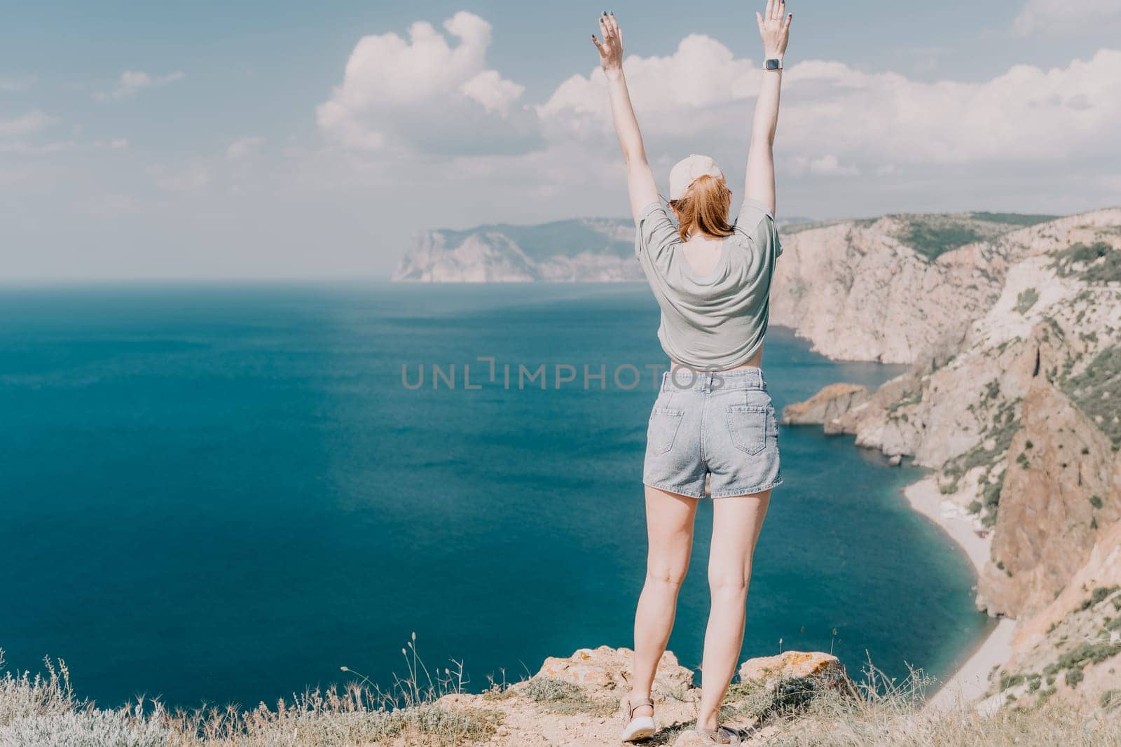 A woman in denim shorts and a green t-shirt stands with arms raised on a cliff overlooking the ocean and distant coast.