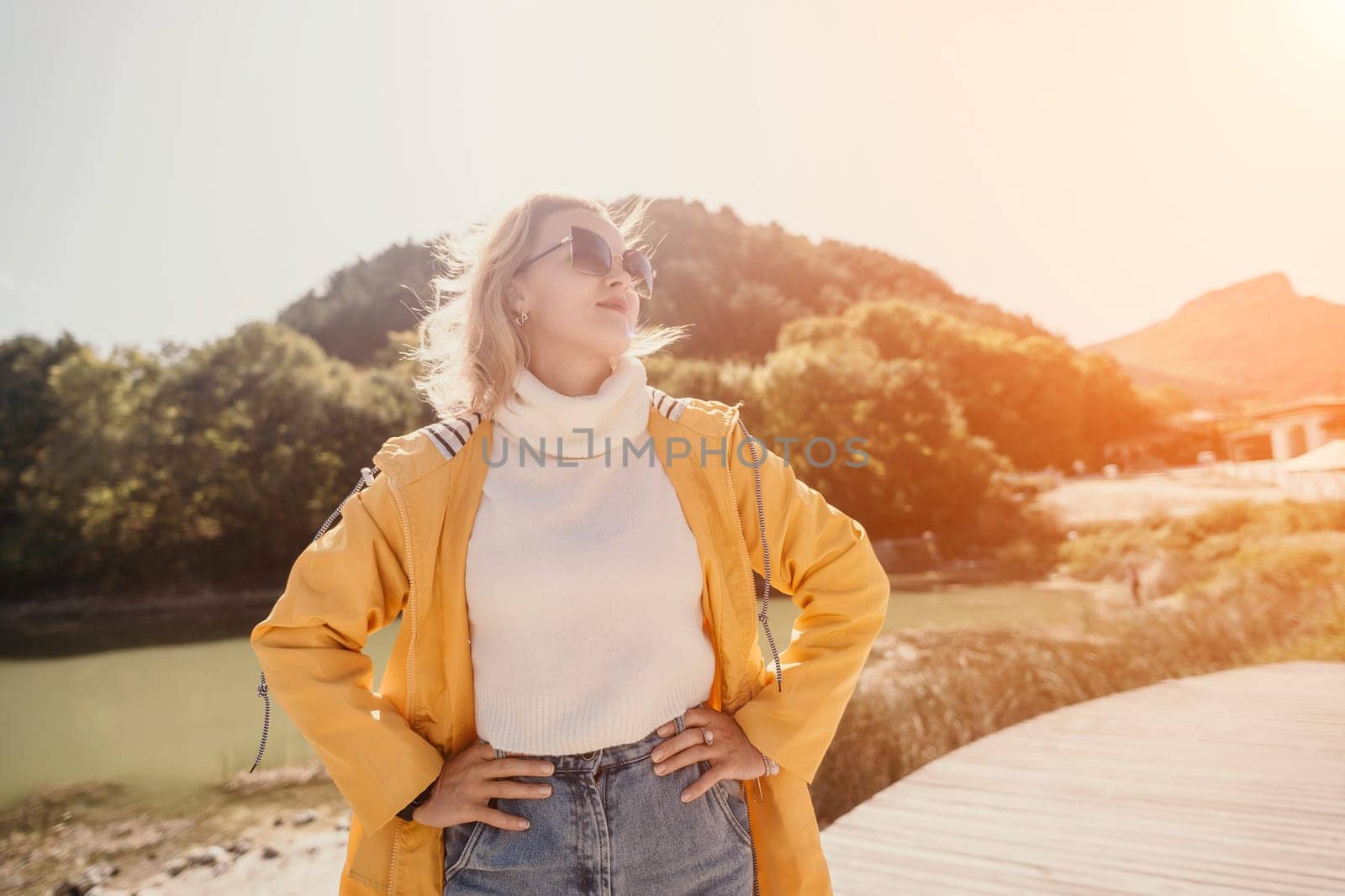A woman in a yellow coat stands with her hands on her hips, looking up at the sun.