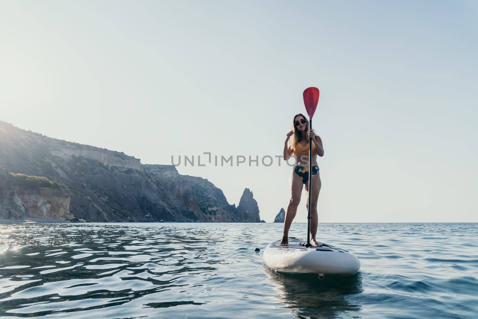 A woman paddles a stand-up paddleboard on calm blue water, with a rocky coastline in the background.