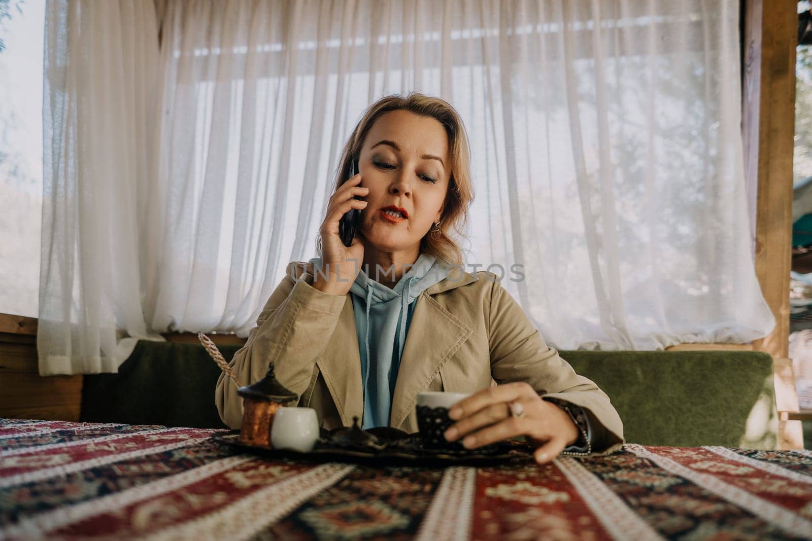 A woman in a beige coat and blue hoodie talks on her phone while sitting at a table in a cafe.