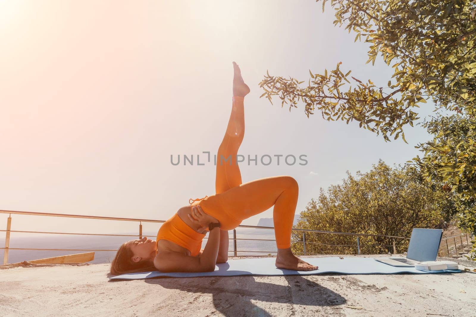 A young woman in an orange yoga outfit performs a challenging pose on a mat overlooking a stunning vista.