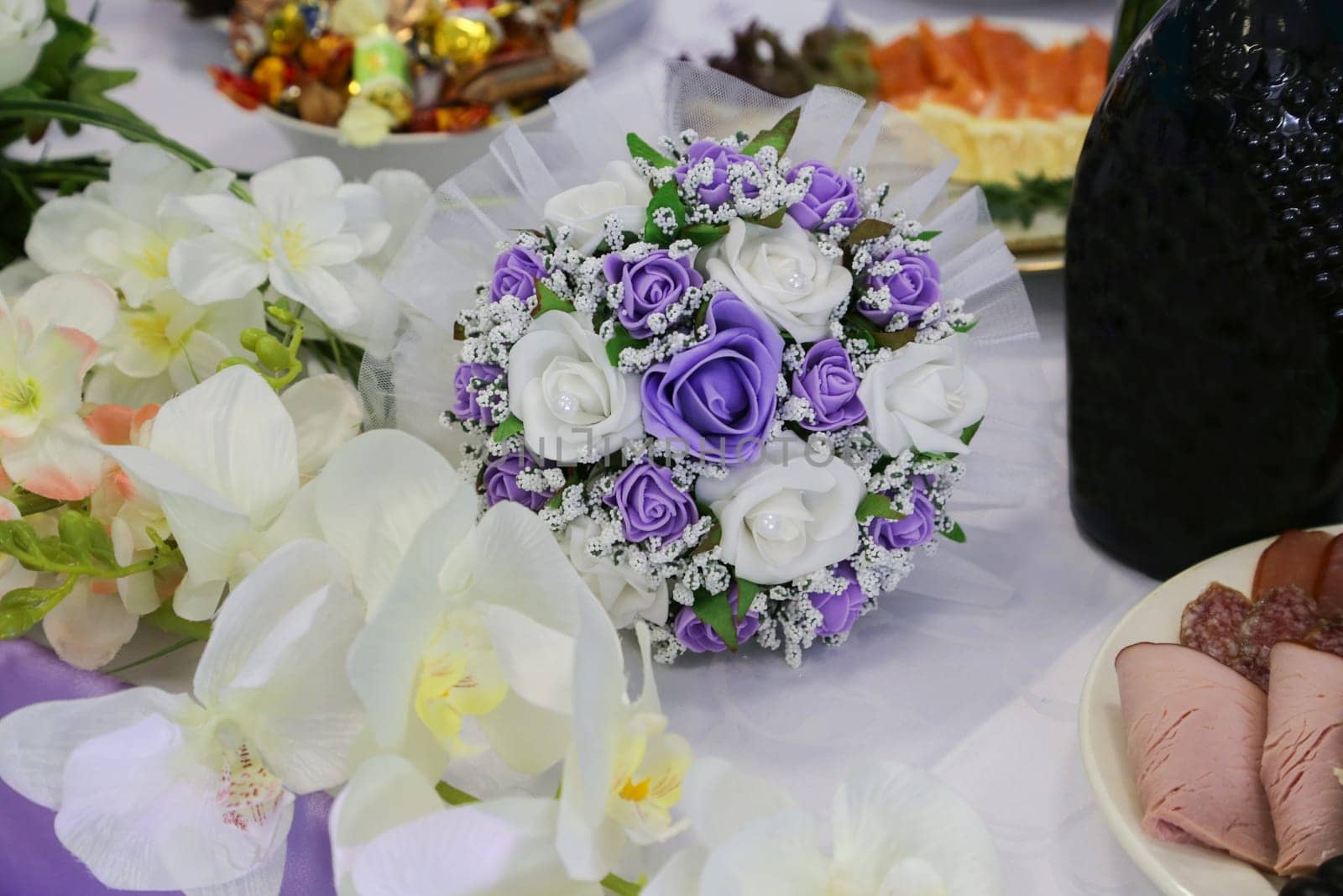 The bride's wedding bouquet with white and lilac flowers lies on the table with festive assorted food and other flowers close-up.