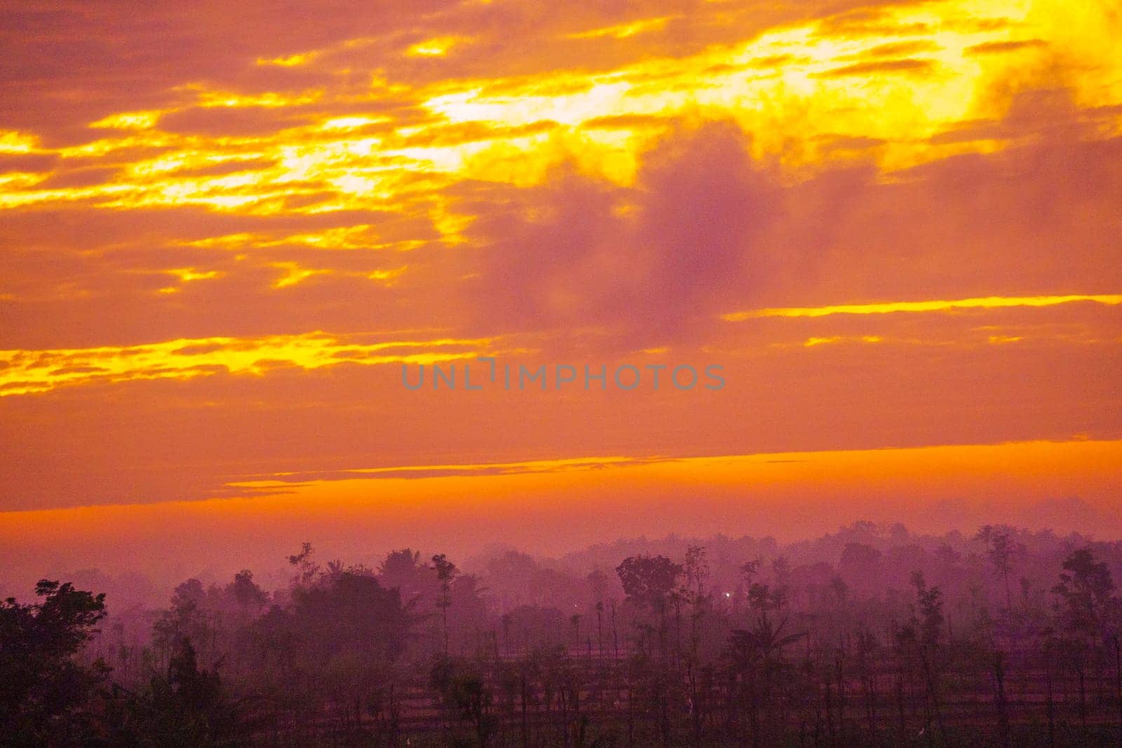 A breathtaking sunrise paints the sky in vibrant hues of orange and pink, casting a warm glow over a rural landscape dotted with trees and fields.