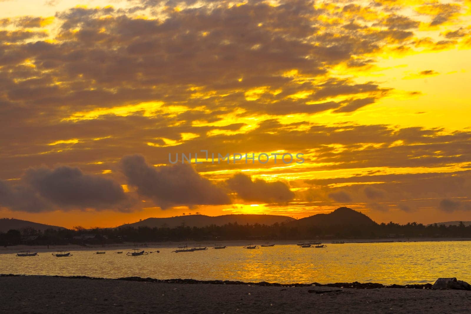 A breathtaking sunrise near Mandalika, Lombok, Indonesia paints the sky in fiery hues over a calm ocean, casting long shadows on the sandy beach and fishing boats.