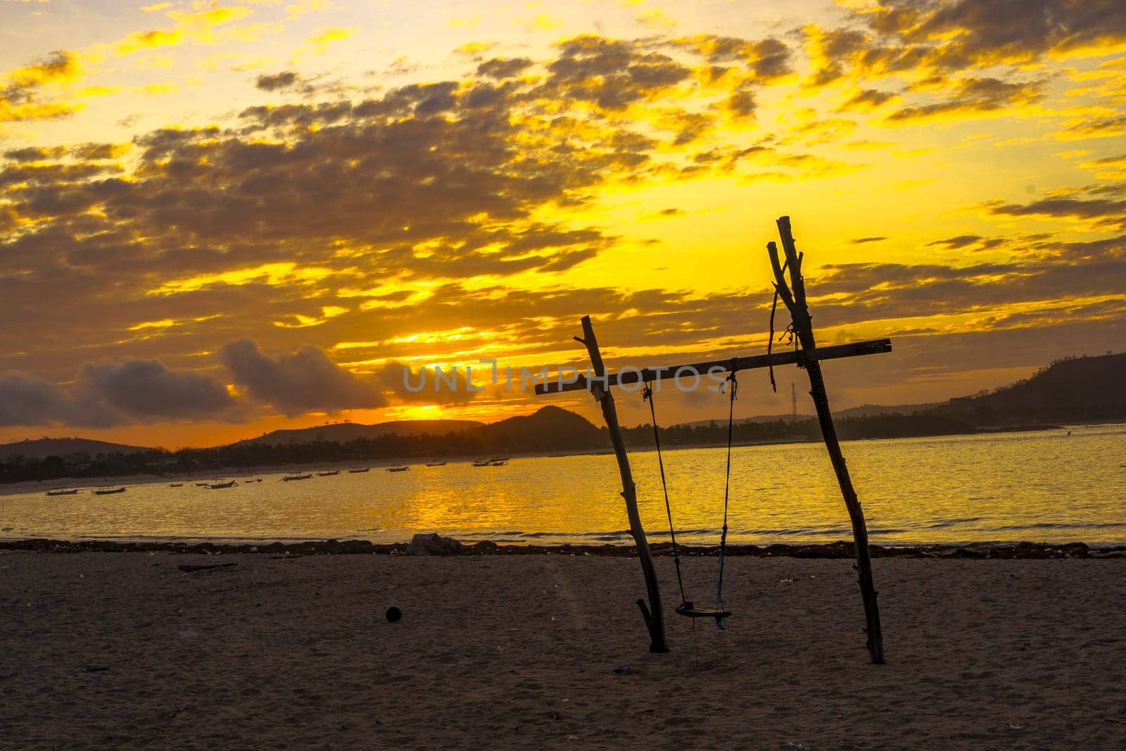 A breathtaking sunrise paints the sky above a secluded beach near Mandalika, Lombok, Indonesia casting a golden glow on the calm waters and highlighting a lone swing on the shore.