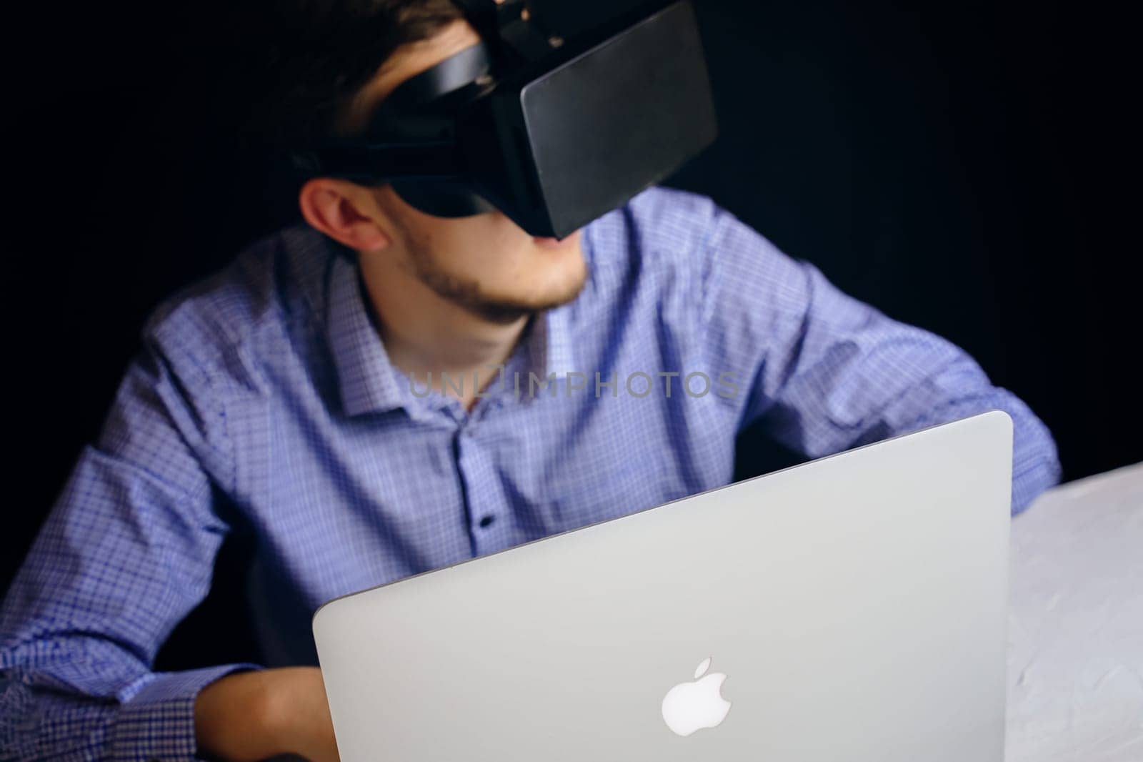 A man is wearing a virtual reality headset while seated in front of an Apple laptop, engaging in a digital experience. Bishkek, Kyrgyzstan - March 14, 2020