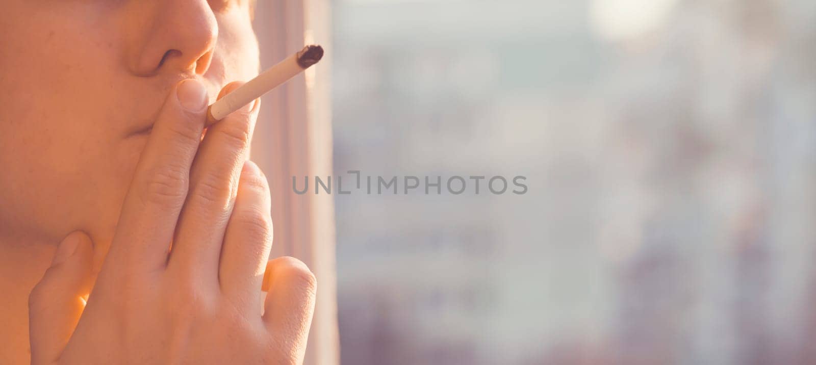 A young man smokes, holds a cigarette in his hand and inhales the smoke while standing at the window early in the morning in the sun, close-up view.