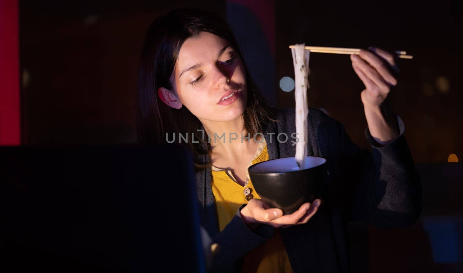 Young girl works at night at home with laptop and eats noodles, asian food with chopsticks. A woman watches a show, a movie and has dinner with ordered food from a cafe.