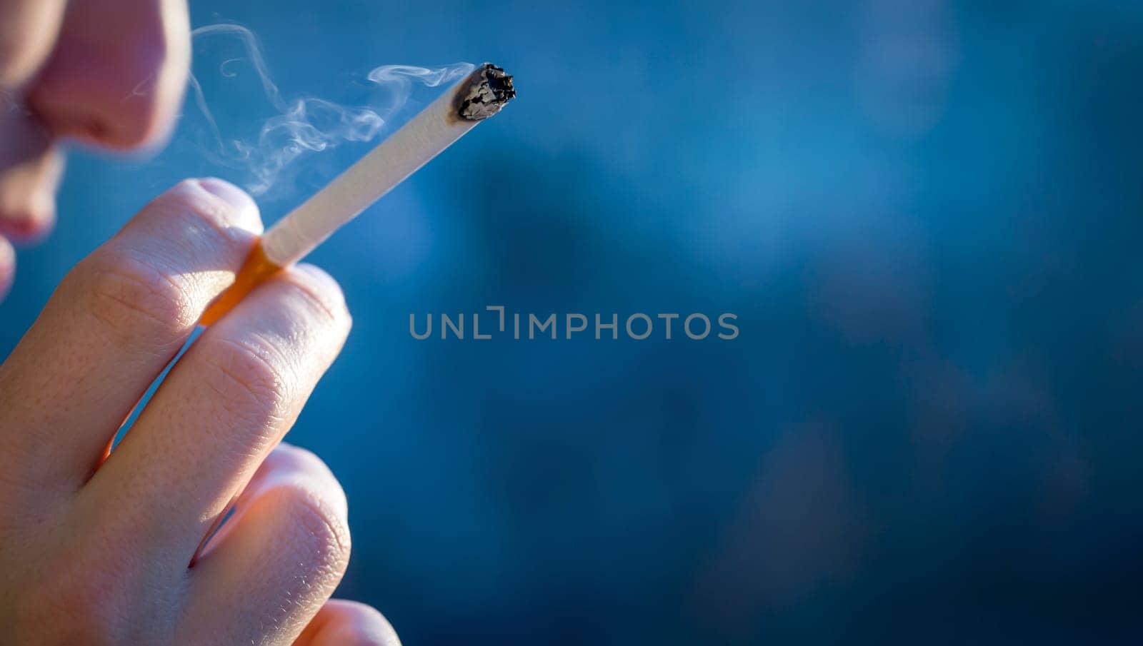 A young man smokes, holds a cigarette in his hand and inhales the smoke while standing at the window early in the morning in the sun, close-up view.