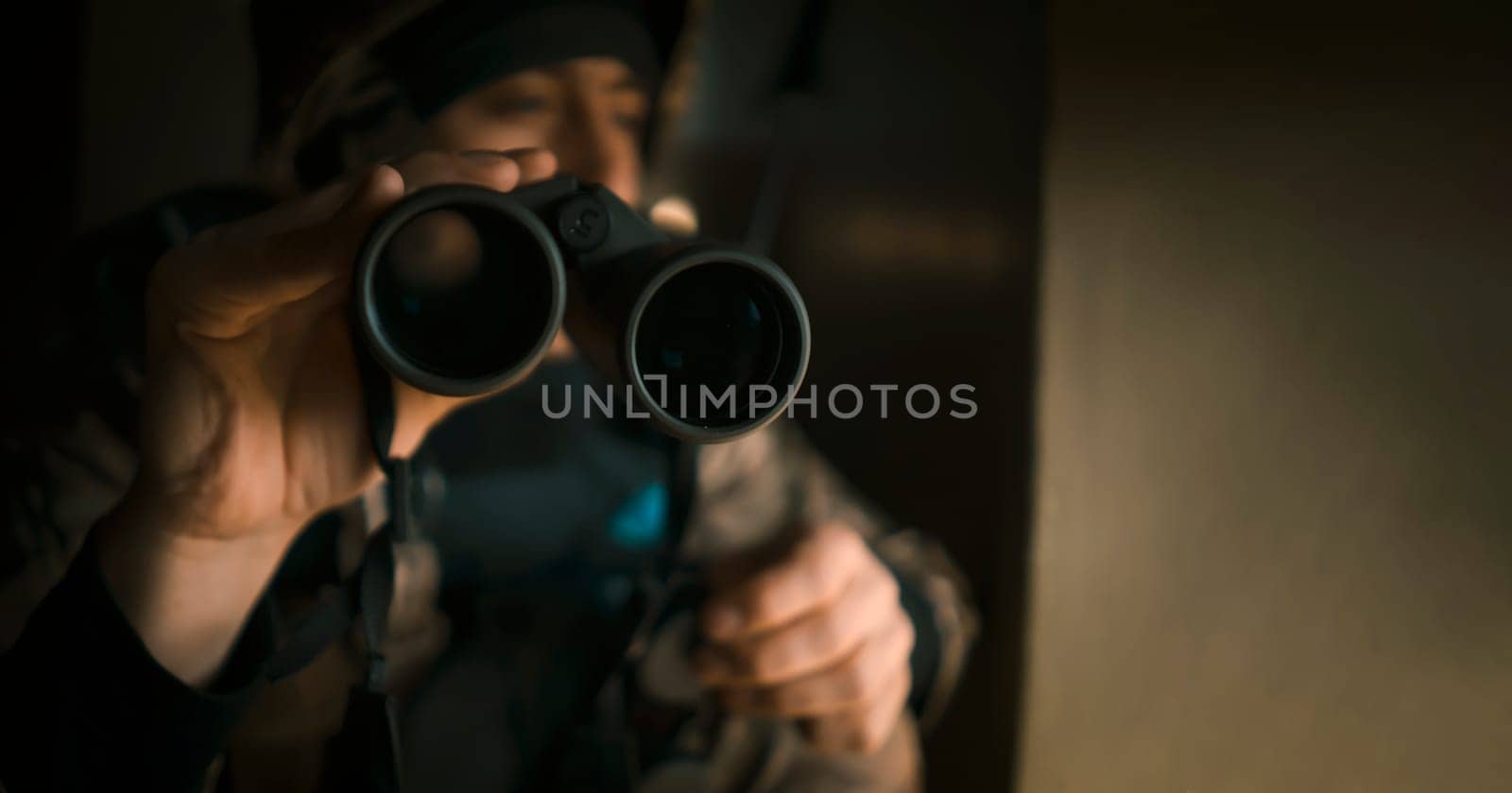 Young man in military uniform and wearing a helmet looks through binoculars during the fighting, undergoes military exercises. The soldier conducts reconnaissance, near the window inside the building.