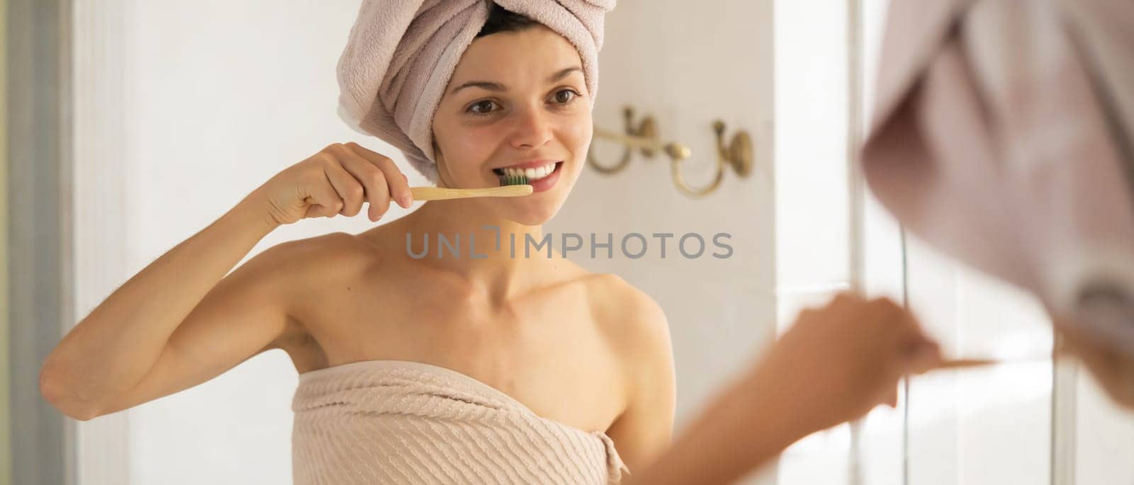 A young girl brushes her teeth near the mirror after relaxing in the bathroom, the woman is wrapped in a towel and takes care of the health and beauty of her body and teeth, gums.