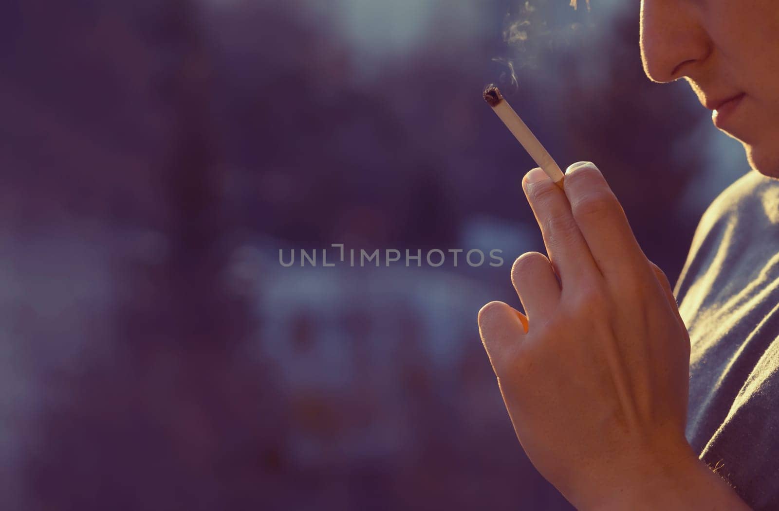 A young man smokes, holds a cigarette in his hand and inhales the smoke while standing at the window early in the morning in the sun, close-up view.