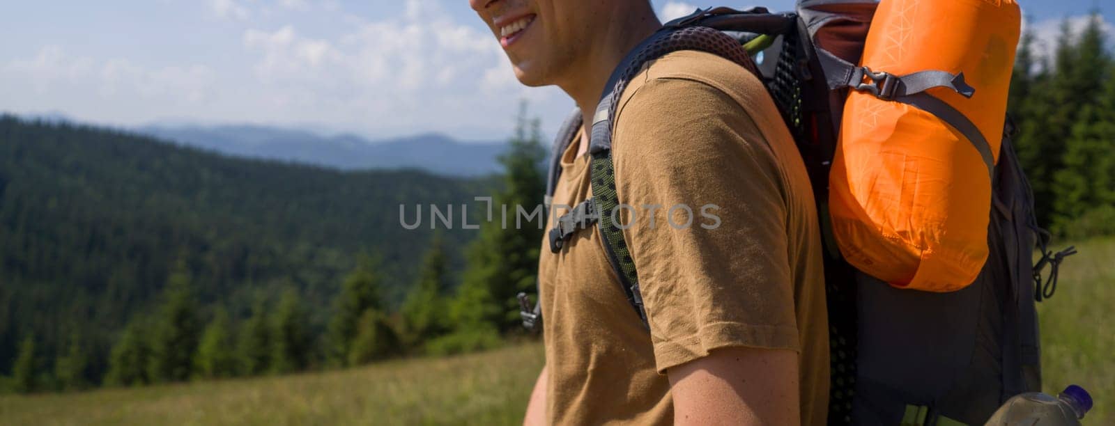 A young man with travel equipment and a large backpack goes on a trip, walks along the road to the national park for hiking in the wild, among the mountains and coniferous forests on a sunny day.