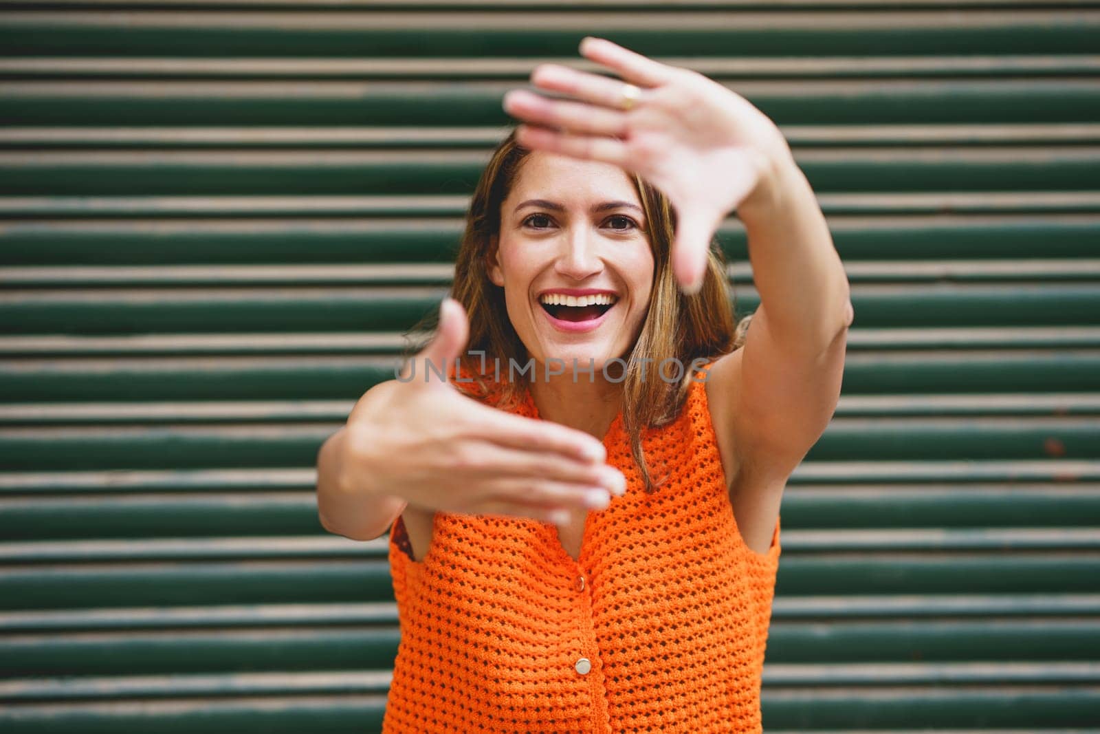 Cheerful latin woman looking at camera and smiling while shaping frame with her fingers in front of wall in city street