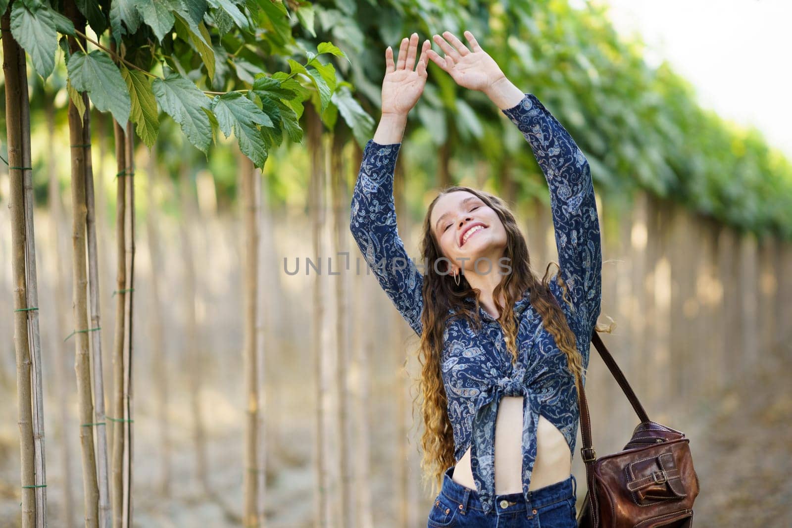 A cheerful young woman enjoys a sunny day in a vineyard, stretching her arms and smiling. The lush green vines create a tranquil atmosphere, highlighting her happiness and sense of freedom