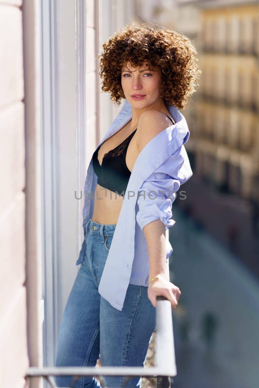 Hot female with brown curly hair in unbuttoned shirt and black bra leaning on railing of balcony looking at camera