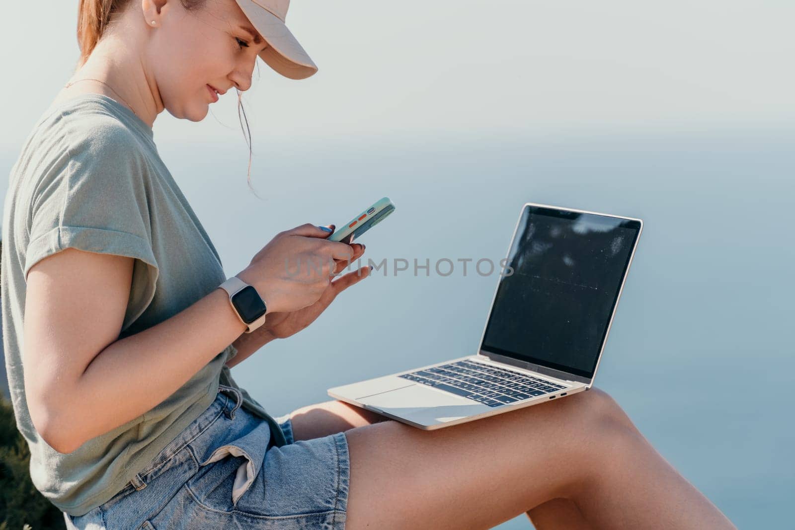 A young woman wearing a baseball cap sits outdoors, checking her phone while a laptop sits on her legs.