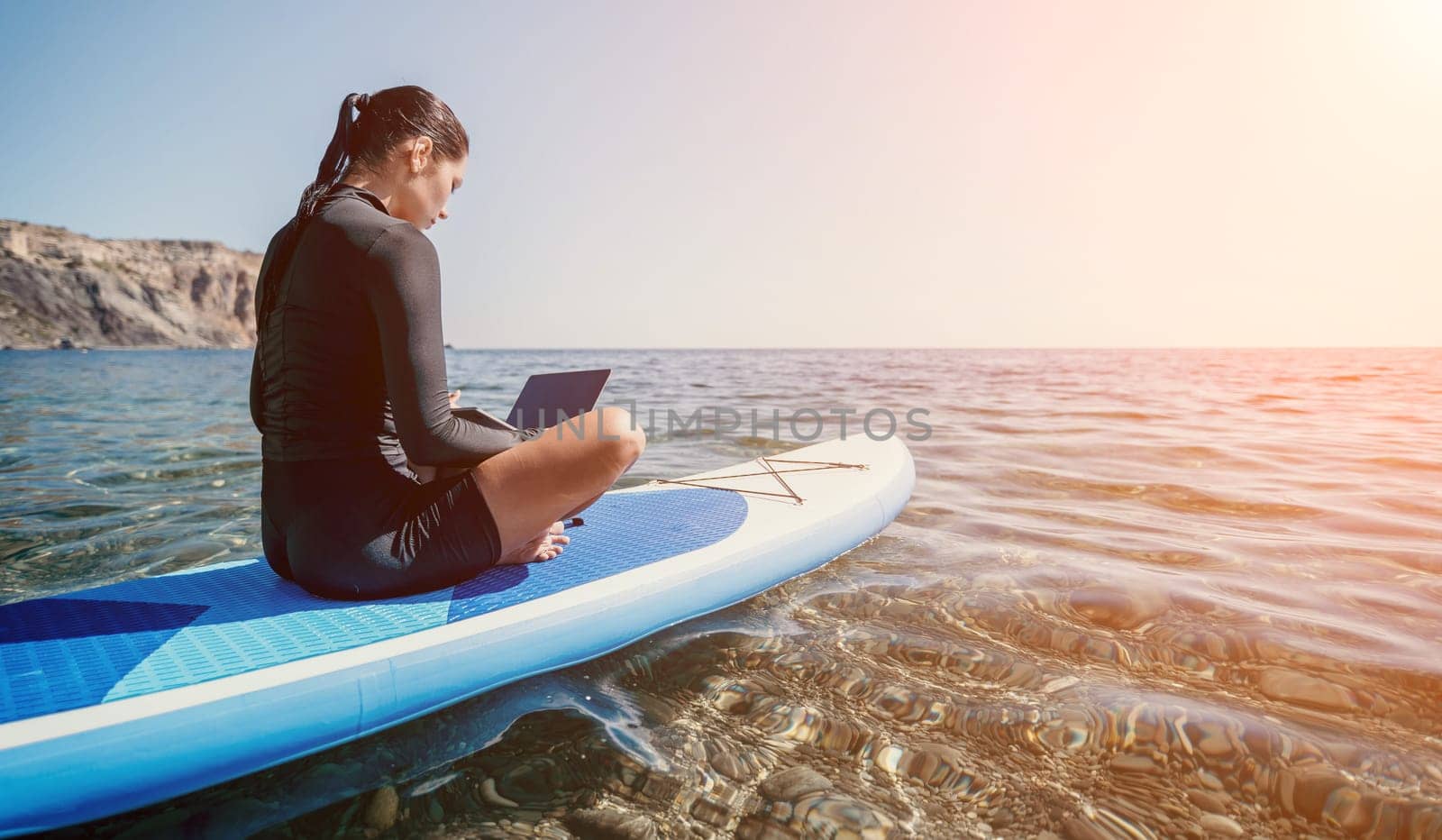 Digital nomad, woman in the hat, a business woman with a laptop sits on the rocks by the sea during sunset, makes a business transaction online from a distance. Freelance, remote work on vacation.