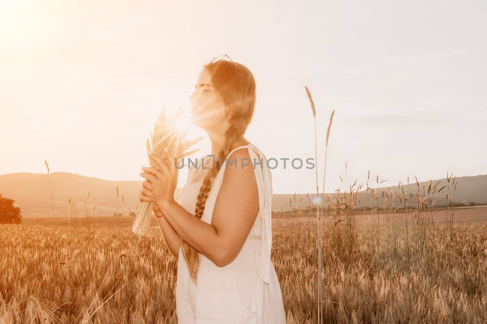 A woman is holding a bunch of wheat in her arms. The wheat is dry and brown, and the woman is wearing a white dress. The scene is set in a field, and the woman is posing for a photo