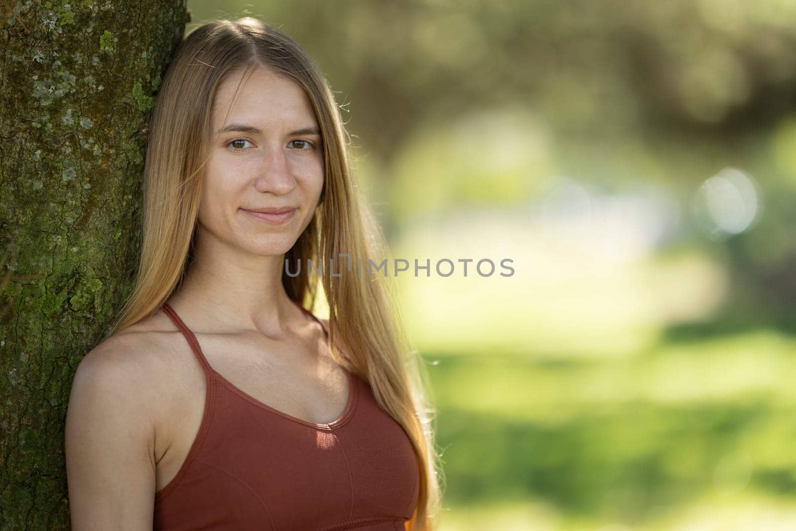 Young woman with long blond hair wearing sportswear, smiling and leaning against a tree in a park