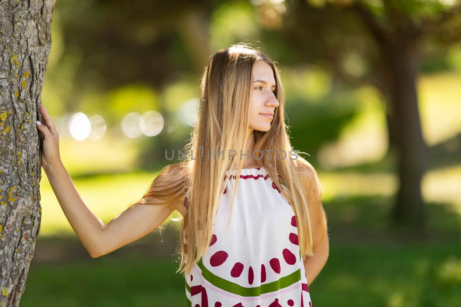 Blonde woman standing next to a tree is taking a peaceful moment in the park