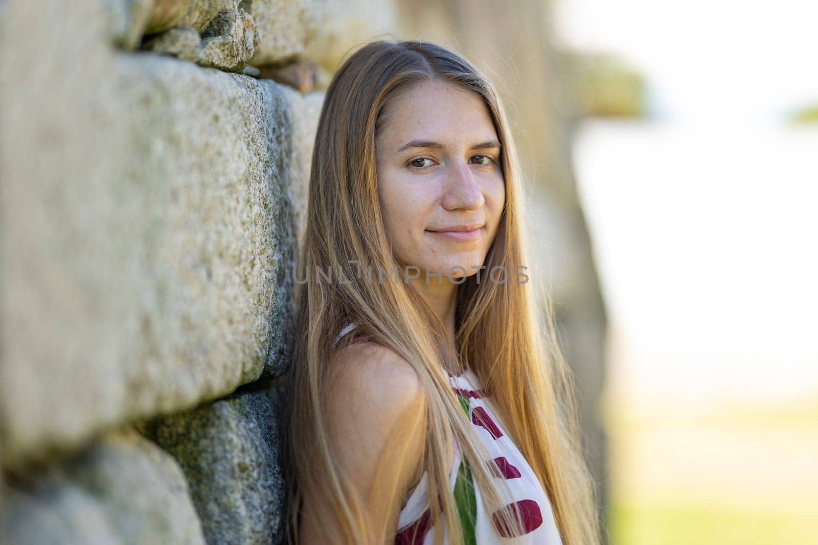 Young blond woman is leaning against a stone wall outside on a sunny day
