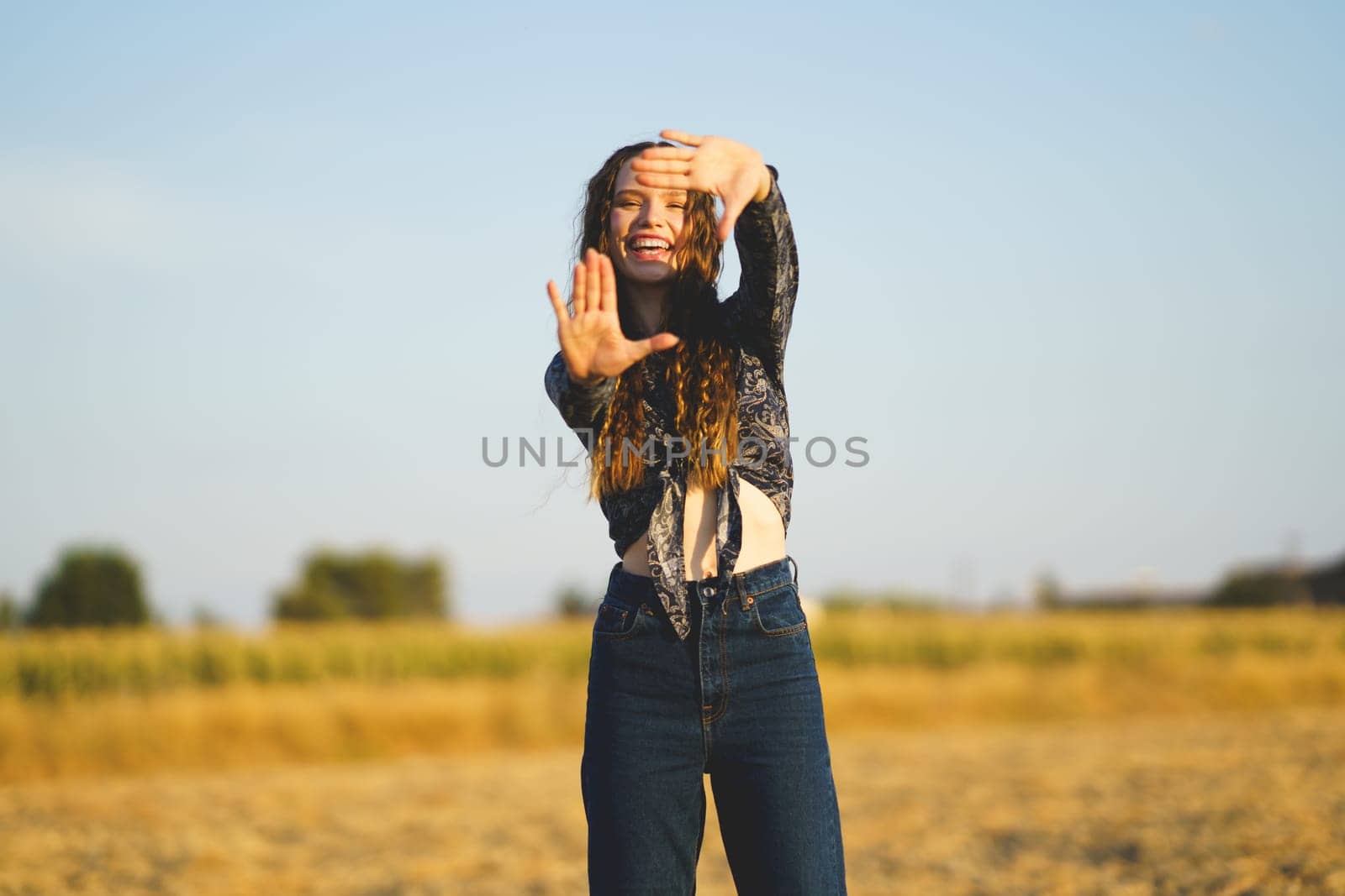 A joyful and enthusiastic young woman is proudly showcasing her creativity while outdoors, striking a playful and lively pose in a beautiful field beneath a clear and bright blue sky