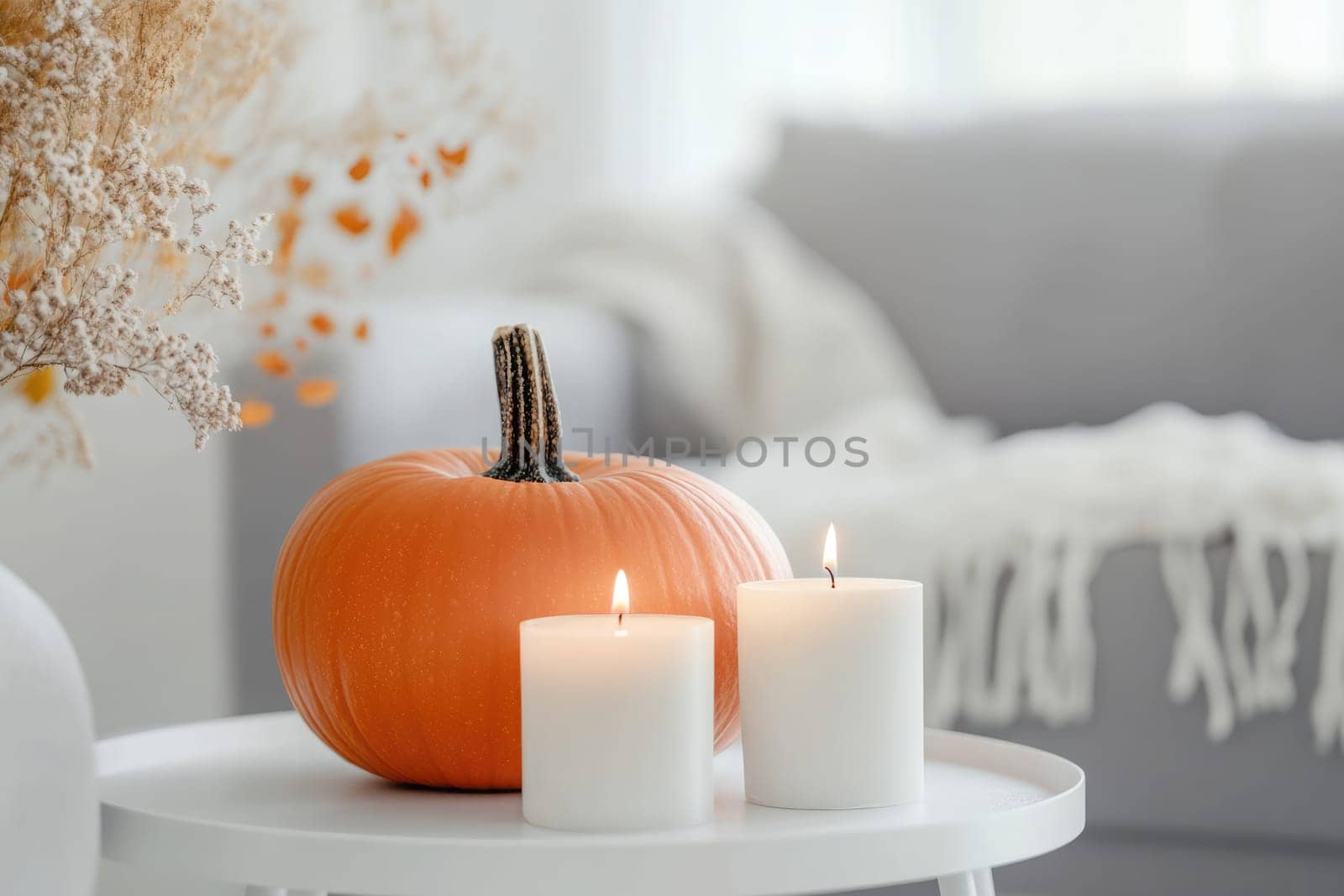 A warm atmosphere fills the living room where a small pumpkin rests next to a lit candle and a vase of dried flowers on a white table, evoking the spirit of autumn.
