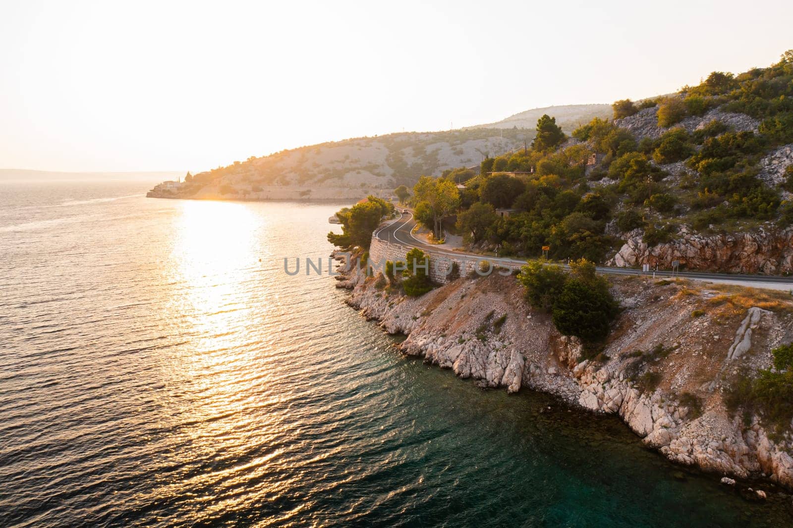 Sunset view of a winding coastal road alongside clear waters in a serene natural landscape.